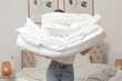 © triocean - Woman holding pile of soft white folded duvet and pillows at home in her bedroom, cozy domestic lifestyle, housewife cleaning, tidying up bedroom, housework chores concept.