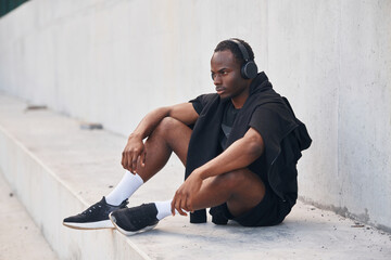 Wall Mural - Sitting by the white wall. Handsome black man is posing outdoors at daytime