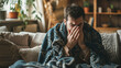Man in blanket with a cold sits on a couch in the living room and blows his nose into a tissue. Cold season flu, coronavirus, winter respiratory infections.
