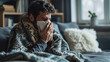 Man with a cold sits on a couch in the living room and sneeze his nose into a tissue. Cold season flu, coronavirus, winter respiratory infections.