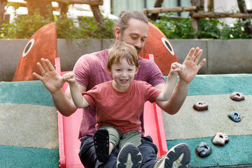 Wall Mural - Caucasian family, father holding his 3 year old son, playing on a slide together in the playground.