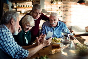 Wall Mural - Group of senior friends enjoying a meal together while using smartphone at home