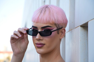 Portrait of young gay boy with pink hair and make-up and wearing pink and black dress and sunglasses is leaning on white wall making different expressions. Concept equality and lgbtq rights.