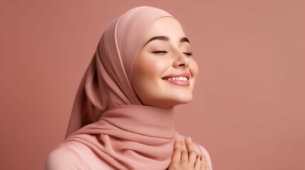 Horizontal shot of positive Muslim woman keeps eyes closed keeps hands on face smiles gladfully wears traditional hijab laughs at something funny isolated on pink background. Positive emotions