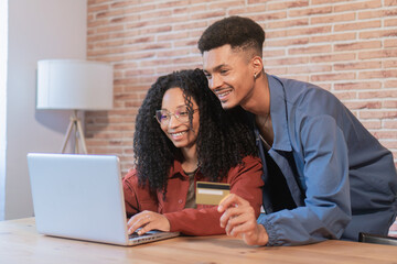 African american young adult couple using the laptop in the living room shopping online with the credit card. Two people boyfriend and girlfriend buying in internet with the pc in a website indoor