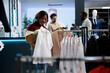 © DC Studio - Clothing store smiling customer holding shirt on hanger and checking apparel fit. Cheerful african american woman examining trying on outfit while shopping in mall boutique