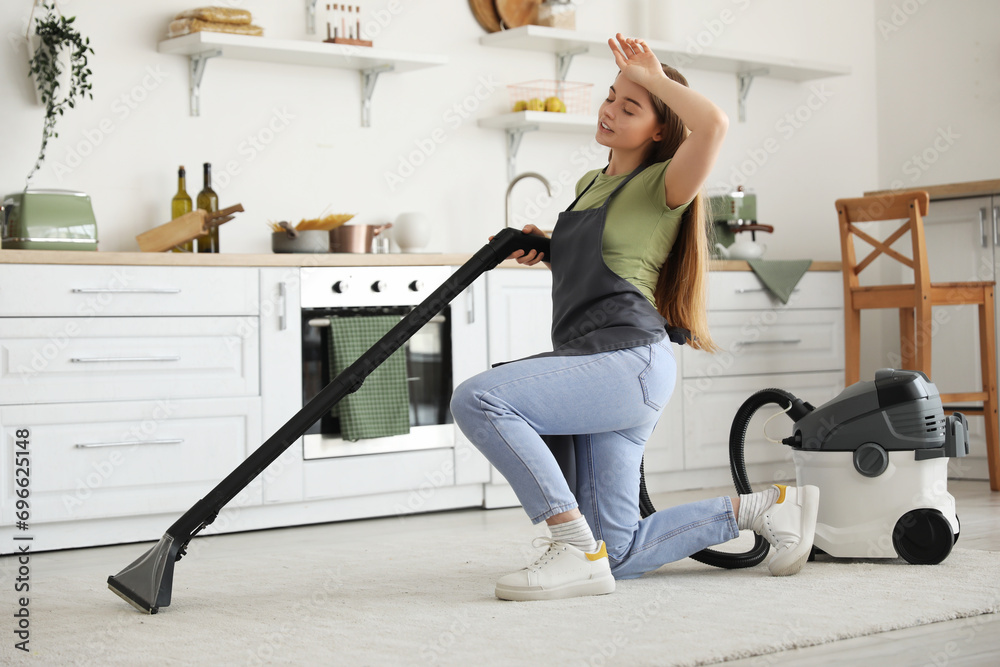 Tired young woman hoovering carpet in kitchen