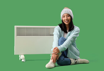 Young Asian woman in winter clothes with radiator sitting on green background