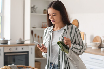 Wall Mural - Young Asian woman with eco bag full of fresh food and mobile phone in kitchen