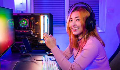 Wall Mural - Smiling Asian woman live stream she play video game via smartphone at home neon lights living room, Gamer playing online game application on mobile phone wear gaming headphones looking to camera