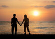 Silhouette lovely Asian couple standing by the sea with beautiful sunset sea and sky background. Back of happy husband and wife holding hands enjoying honeymoon relaxing on tropical summer vacation.