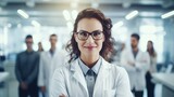 Fototapeta Nowy Jork - Beautiful young woman scientist wearing white coat and glasses in modern Medical Science Laboratory with Team of Specialists on background. copy space for text.