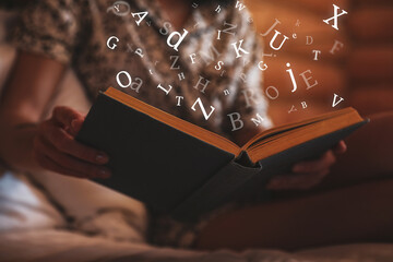 Wall Mural - Woman reading book with letters flying over it on bed at home, closeup