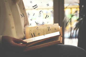 Wall Mural - Woman reading book with letters flying over it indoors, closeup