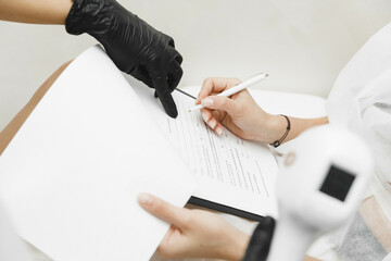 female client signs consent form while sitting during medical consultation with cosmetologist. Compliance with legal requirements when receiving personalized medical care. hair removal