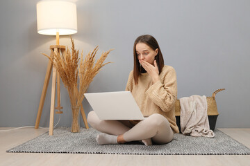 Wall Mural - Remote job. Online freelance opportunities. Shocked Caucasian woman wearing beige jumper working on computer having troubles with saving information while sitting on floor in light room