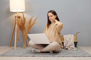 Wall Mural - Freelance flexibility. Remote work. Tired overworked brown haired woman wearing beige sweater working on computer having pain in neck while sitting on floor in home interior