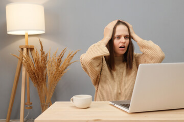 Wall Mural - Networking workspace professional. Despair shocked sad brown haired woman wearing beige sweater working on computer while sitting at table in home office keeps hand on head