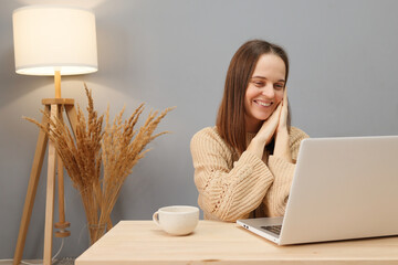 Wall Mural - Laptop for remote work. Attractive pretty brown haired woman wearing beige sweater working on computer looking at screen with cheerful smile while sitting at table against lamp and dry flowers