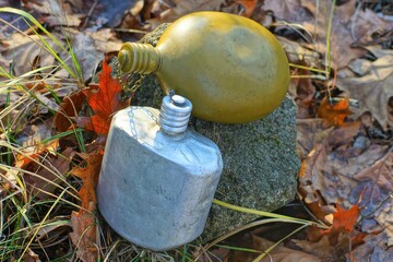 two different empty dirty  old  manual aluminum gray and olive   army  closed flasks lie on the ground near a gray stone  on the street 
