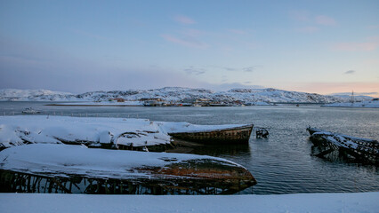 Sticker - Snow-covered abandoned boats on a frozen shore with a backdrop of a snowy village at twilight, conveying desolation in a winter setting