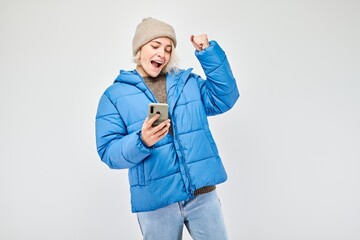 Wall Mural - Portrait of young blond woman in blue jacket with mobile phone in hand celebrates victory, winning money in casino. Person with smartphone isolated on white background.