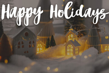 Wall Mural - Happy Holidays text on cozy christmas miniature village with glowing lights in evening. Merry Christmas. Season's greeting card. Handwritten sign