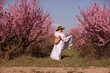 Woman blooming peach orchard. Against the backdrop of a picturesque peach orchard, a woman in a long white dress and hat enjoys a peaceful walk in the park, surrounded by the beauty of nature.