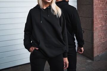 Wall Mural - A mock-up template of black hoodies worn by a couple. A design concept for print and branding. A couple poses outdoors in a casual and elegant street wear style. No face and no logo shown.