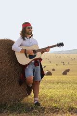 Wall Mural - Hippie man playing guitar near hay bale in field