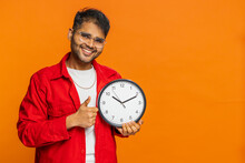 It is your time, hurry up. Indian young man showing time on wall office clock, ok, thumb up approve pointing finger at camera, advertisement, deadline. Arabian guy on orange background. Copy-space