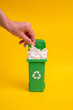 hand puts garbage in trash can with paper on yellow background