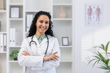 Portrait of young beautiful successful female doctor inside medical office, hispanic woman with curly hair in white medical coat smiling and looking at camera with arms crossed, doctor inside clinic.