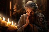 Fototapeta  - An elderly woman with gray hair prays with her hands up to her face near burning candles in a church