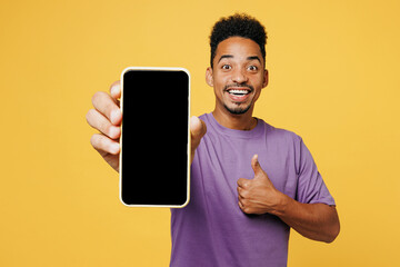 Wall Mural - Young man of African American ethnicity wear purple t-shirt casual clothes hold use close up blank screen area mobile cell phone show thumb up isolated on plain yellow background. Lifestyle concept.