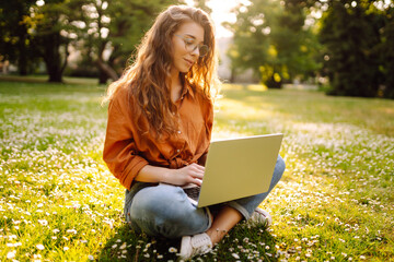 Wall Mural - Portrait of a happy woman in a sunny park on a green lawn with a laptop. Young freelancer woman working on a laptop, enjoying the sunny weather. Freelance concept, technology.