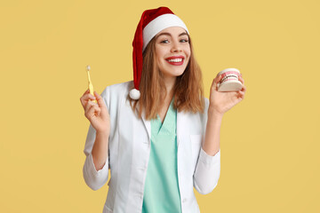 Canvas Print - Young female dentist in Santa hat with toothbrush and jaw model on yellow background