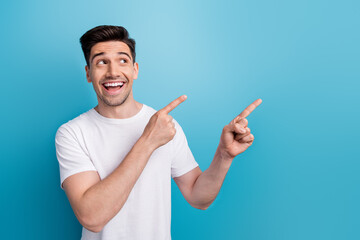 Wall Mural - Photo of excited dreamy man dressed white t-shirt pointing two fingers looking empty space isolated blue color background