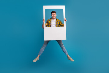 Poster - Full body shooting young funny guy jumping happy smile holding portrait frame during photo session isolated on blue color background