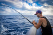 Tough man wearing baseball cap on board ship while fishing against blue sea background.