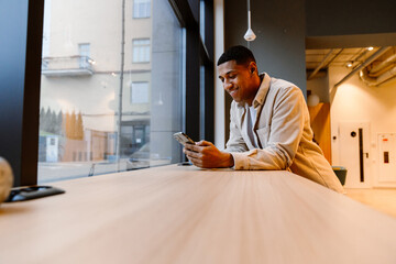 Wall Mural - Smiling african man using smartphone online mobile app while standing in office