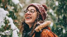 Laughing Young Woman In A Knitted Hat, Jacket And Glasses Is Happy To See The First Snow On A Walk. In The Background Snowy Forest With Christmas Trees, Morning.