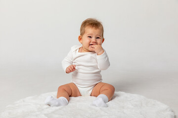 Wall Mural - Happy smiling adorable newborn baby girl in white bodysuit sitting on light white home carpet on white background. Posing and looking at camera. 6 to 7 months old infant development.