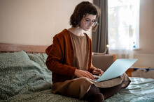 Busy thoughtful teenage girl sitting on bed with laptop computer, thinking, reading text document, doing homework while studying online, working as freelancer, blogger, writing book in home atmosphere