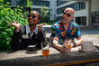 Fashionable mid-age man carrying a colorful hawaiian shirt and actractive young woman of color dressed in black sit at a bar table with drinks and smile enjoying the conversation in a bright sunny day