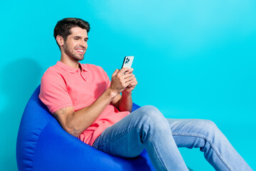 Poster - Photo of cheerful relaxed young guy sitting bean bag browsing social media enjoying twitter posts isolated on aquamarine color background