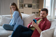 Frustrated jealous woman looking angry at husband who reading messages in phone sitting on couch at home. Husband ignoring wife. Betrayal infidelity mistrust misunderstanding marital discord concept.