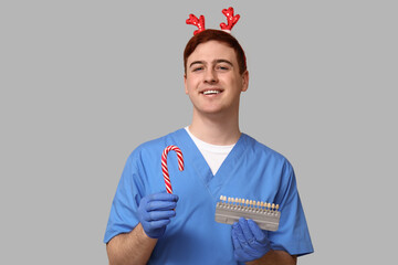 Sticker - Portrait of young male dentist in Christmas reindeer headband with candy cane and dental shade guide on grey background