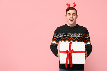 Poster - Handsome young man in warm sweater and reindeer headband holding gift box on pink background