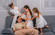 © dvulikaia - Exhausted young father sits on the couch while their four daughters run around and scream. Difficulties of large family concept.